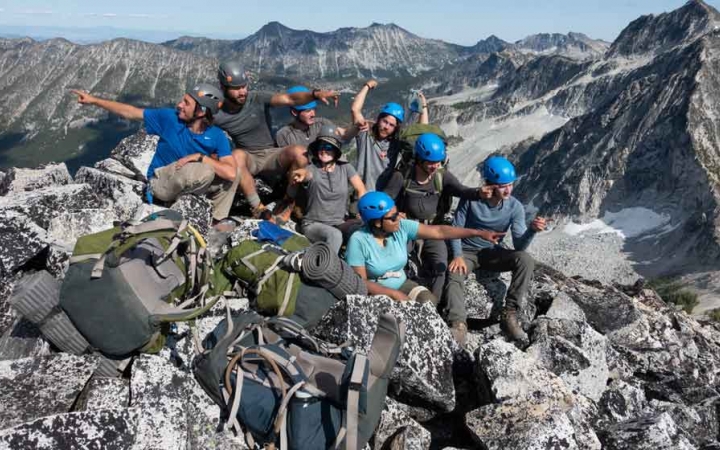 a group of gap year students sit atop a summit and make silly poses. there are mountains in the background